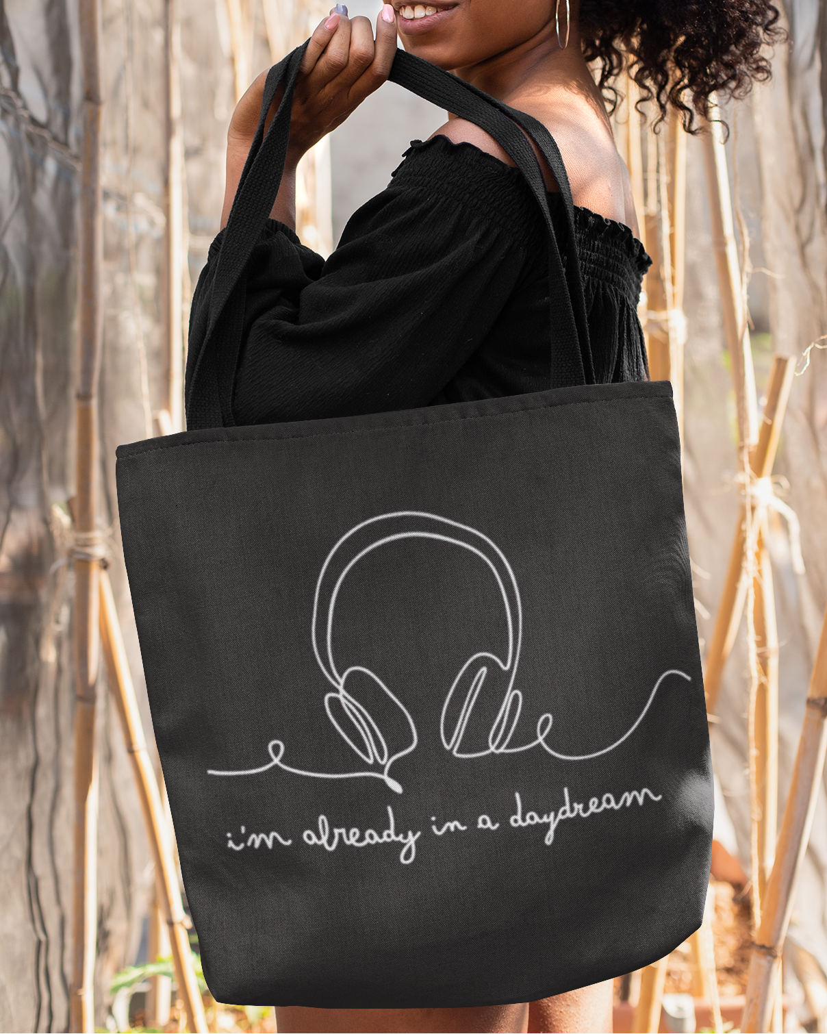 Woman holding a black tote bag with headphones design and text in a natural setting