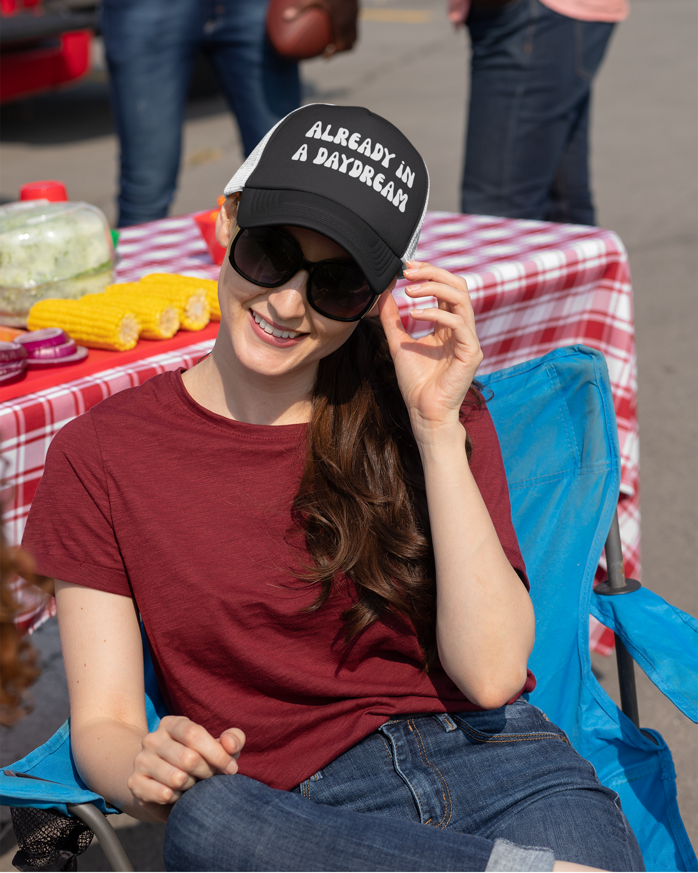 Woman wearing a black cap with text, sunglasses, and red shirt sitting at a table outdoors.