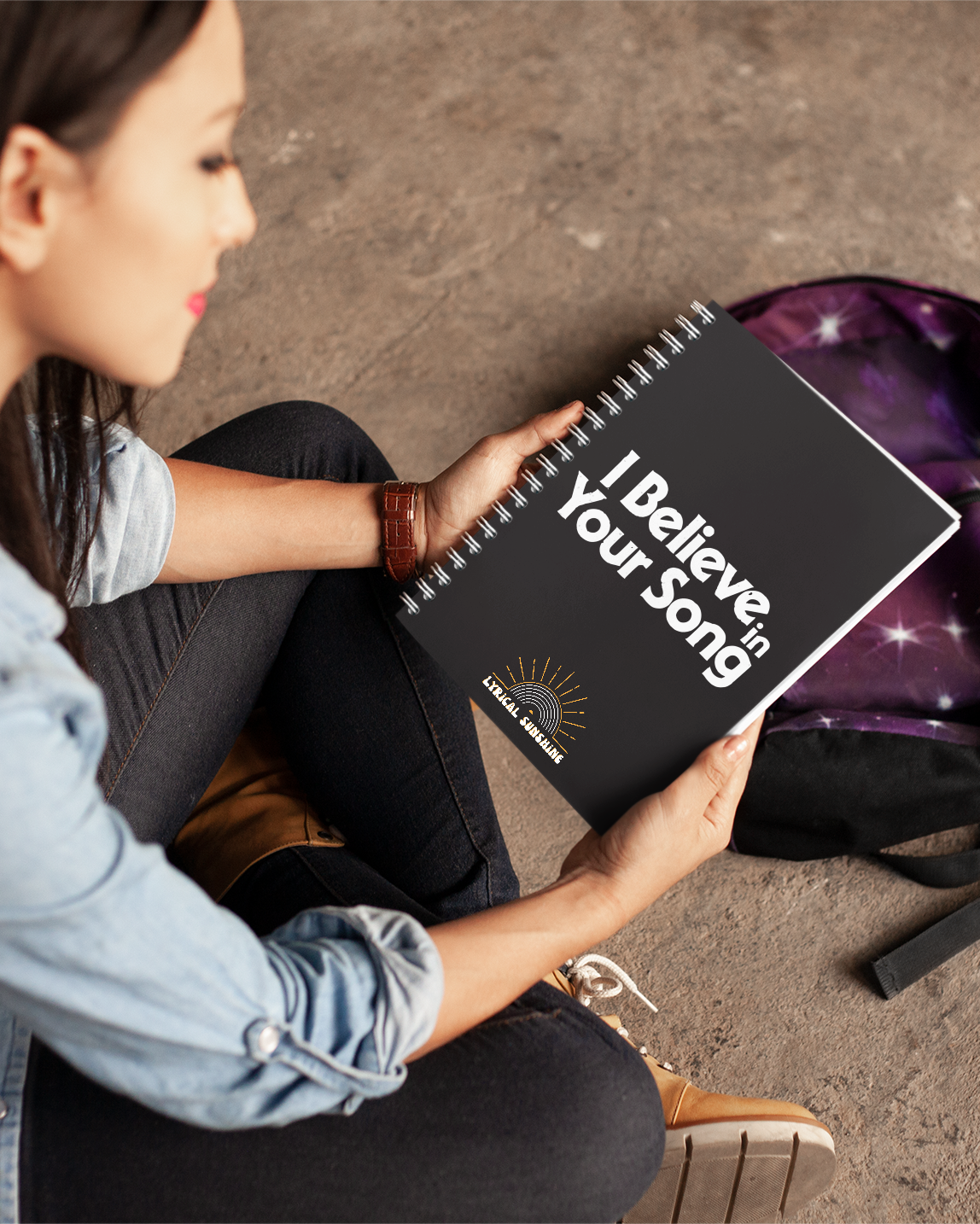 Woman holding a black notebook titled 'I Believe Your Song' with a purple backpack in the background.
